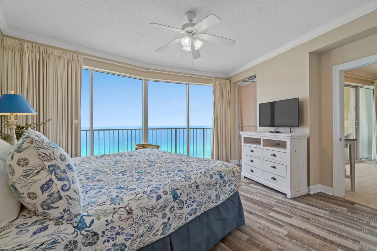 A second bedroom with floral bedding, large windows revealing ocean views, and a TV on the dresser. The room is bright and peaceful, perfect for children, friends, or grandparents. The setting encourages both relaxation and entertainment.