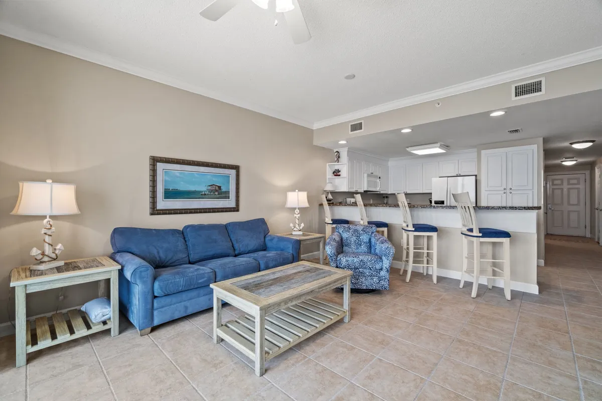 The photo presents a welcoming living area with a blue sofa, coffee table, and bar stools under colorful, beach-inspired artwork. The space is arranged for relaxation, socializing, or unwinding after a day at the beach. It’s an inviting scene that signals the condo’s thoughtful design for groups and families.