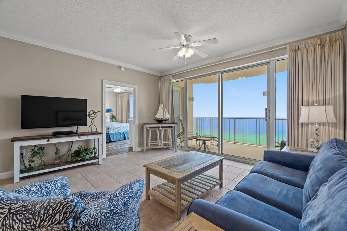 This image features a spacious living room with a blue sofa, large sliding glass doors that open to an ocean view, a wooden coffee table, and a television. The sunlight pours into the room, highlighting the comfortable seating and the seamless connection to the outdoors.
