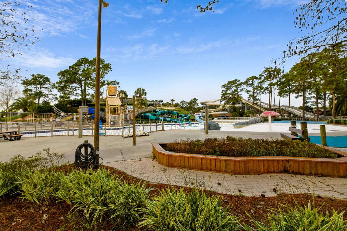 This vibrant exterior scene captures a lively waterpark area with twisting water slides, a sparkling swimming pool, landscaped gardens, and a picnic area. Families and children can be seen enjoying the water activities and gathering for outdoor fun under the sun.
