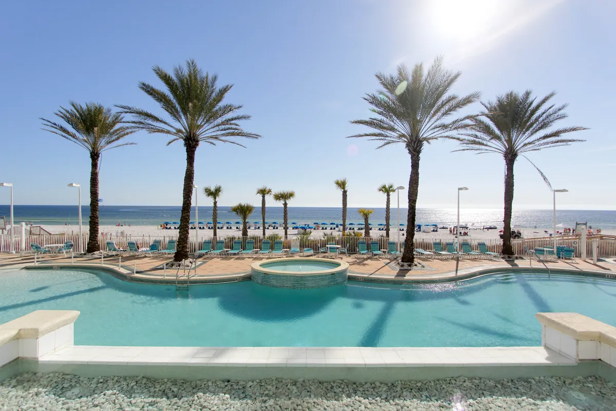 A large, curved swimming pool with an integrated hot tub sits in the foreground, flanked by rows of lounge chairs and shaded by tall palm trees. Beyond the pool, the sandy beach and calm Gulf waters stretch out, with organized chairs and umbrellas signaling easy beach access. The whole area exudes a relaxed, tropical resort vibe.