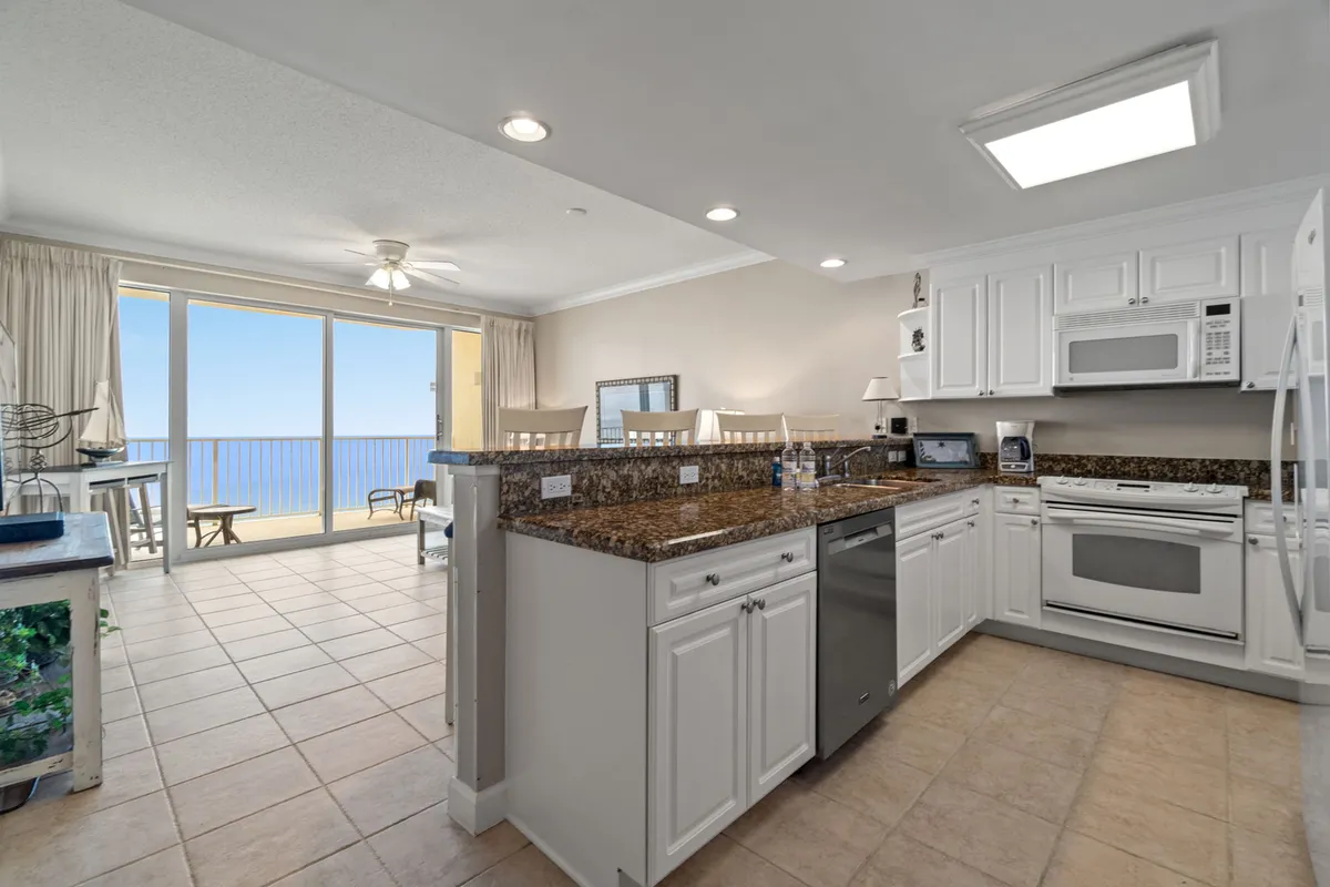 A bright, airy kitchen with a granite countertop island, white cabinetry, full-size appliances, and bar stools. The kitchen is open to the living area and features a direct view through sliding glass doors to the balcony and the Gulf coast beyond. The laundry area is visible adjacent to the kitchen, emphasizing the ease and practicality of the space.