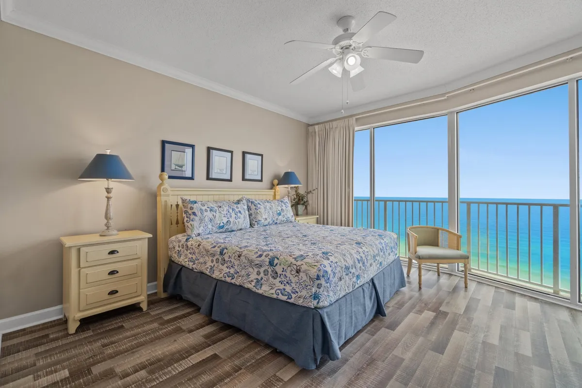 This image showcases a spacious primary bedroom with a large king-size bed adorned in floral bedding. Floor-to-ceiling windows offer a stunning Gulf Coast ocean view, and a private balcony with seating overlooks the beach. Coastal-themed artwork and bedside lamps enhance the tranquil ambiance.