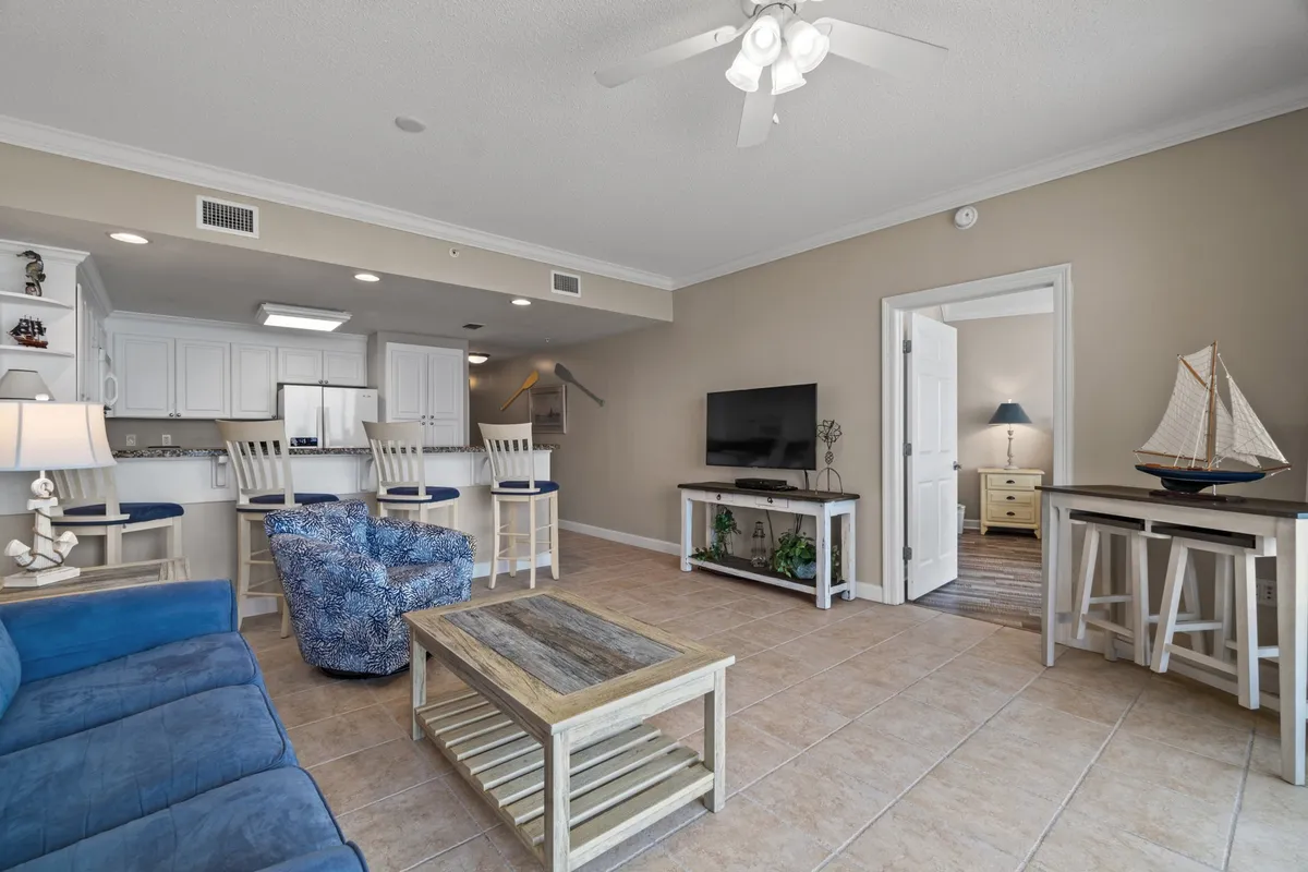 The image displays a bright, open living room with a blue sofa adorned with nautical-themed cushions, a rustic wooden coffee table, and a white kitchen with bar stools in the background. Decorative nautical elements, such as a model sailboat and oars, reinforce the coastal ambiance. Large sliding glass doors offer a glimpse of the balcony and invite natural light into the space.