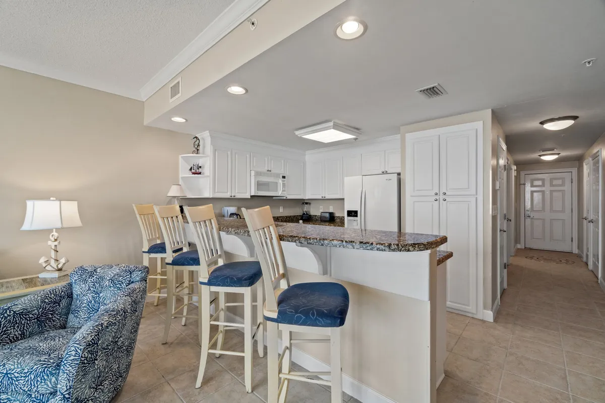 This image showcases an open kitchen layout with a granite countertop and bar stools, white cabinetry and appliances, and a hallway leading to the bedrooms. The kitchen area is bright and welcoming, inviting casual gatherings and easy meal preparation.