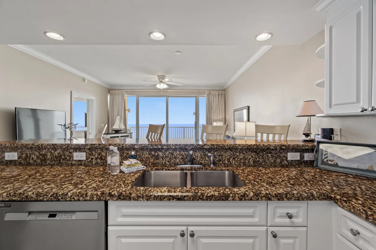 A bright dining area adjacent to a kitchen with granite countertops and nautical decorative items. Large windows provide floor-to-ceiling Gulf views, flooding the space with natural light and highlighting the open-concept layout. The table and counter seating invite both casual and formal dining experiences.