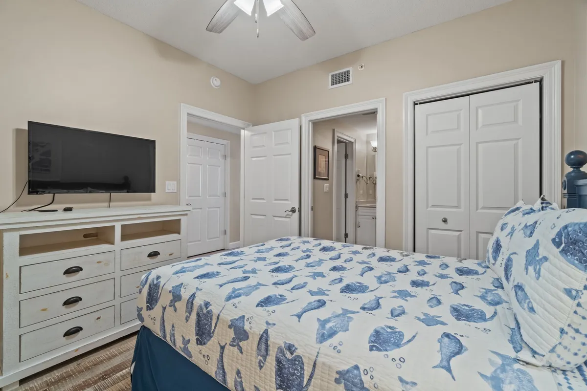 A cozy bedroom with a fish-patterned quilt and white dresser, including a mounted TV. The open door reveals a connected bathroom, and the room is decorated in a nautical theme with soft, relaxing hues.
