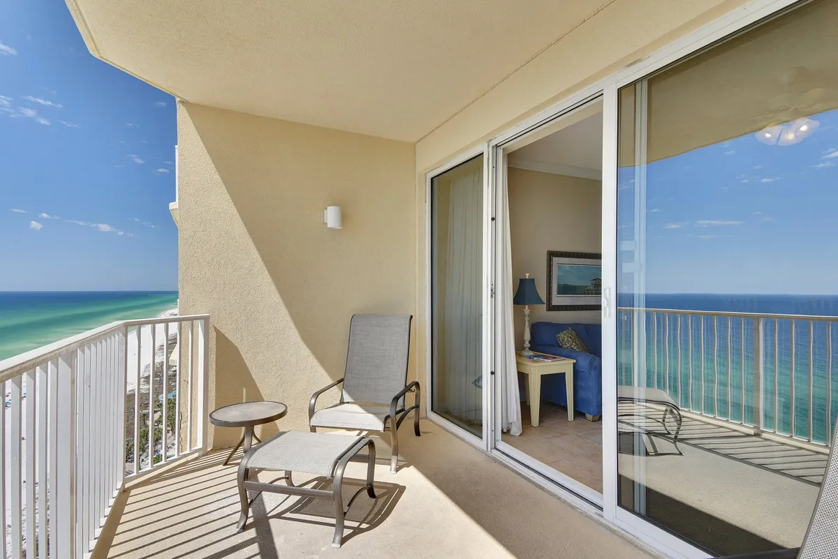 A cozy balcony setting with a comfortable chair and ottoman, positioned for relaxation and taking in the expansive ocean view. The sliding glass doors reflect the water, and the interior blue sofa is visible, signaling the easy flow between indoor dining and outdoor enjoyment.