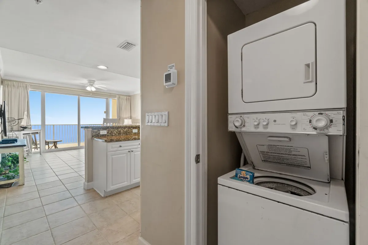 A kitchen and dining space with a granite island, stacked washer and dryer, and floor-to-ceiling balcony windows framing a beautiful ocean view. Nautical-themed décor adds a beachy touch to the inviting and functional space, and the open-plan design keeps the kitchen, dining, and living areas connected.