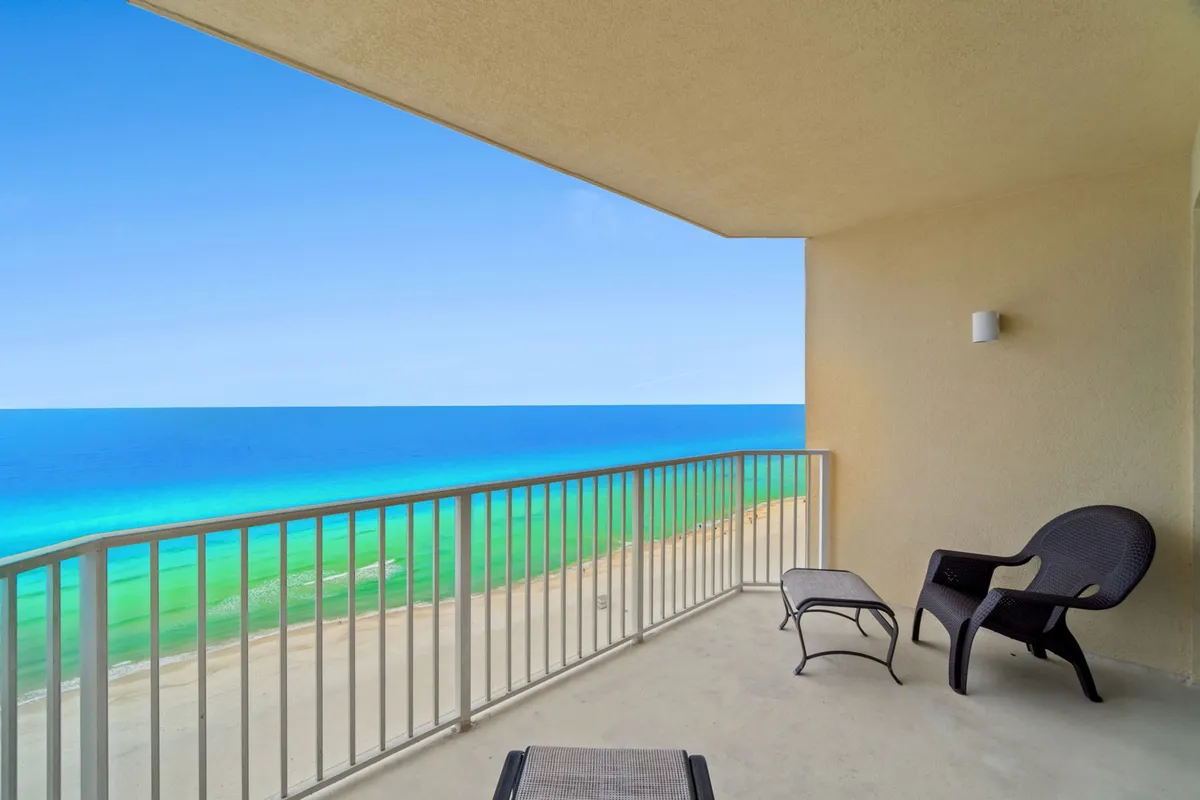This image features a private balcony overlooking the ocean, with a black lounge chair and footrest positioned to enjoy uninterrupted views of the turquoise Gulf waters and endless sandy beach below. The scene is bathed in calming natural light, offering a sense of peaceful seclusion and relaxation.