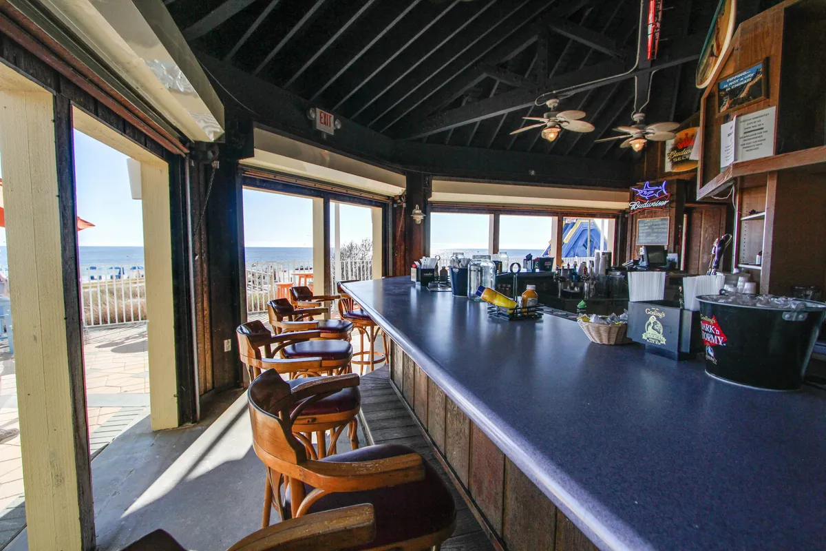 A long bar counter with stools is placed beside an open wall/window, offering stunning beach and ocean views. The area features a wooden ceiling with exposed beams, nautical decor, and a neon sign, creating a lively, coastal atmosphere.