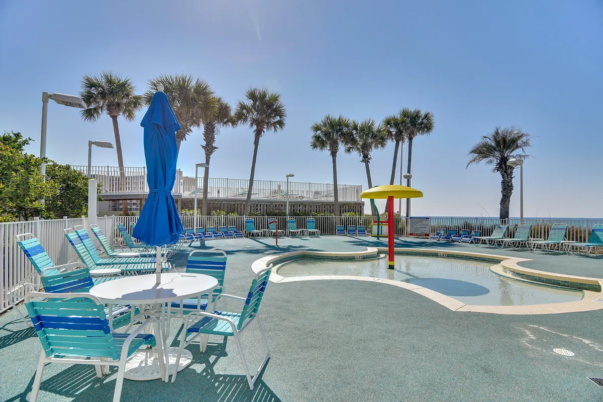 A kid-friendly splash zone pool with a water mushroom feature is surrounded by poolside lounge chairs with blue and white stripes. Palm trees line the background, and a white round table with a blue umbrella adds to the resort feel.