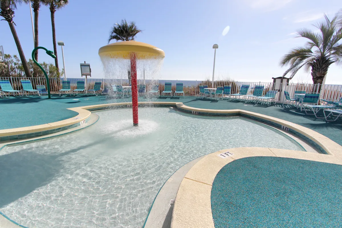 A vibrant children's splash zone water feature with a cascading water umbrella sits at the center, surrounded by turquoise lounge chairs and palm trees. The ocean horizon is visible in the background, highlighting the proximity to the beach.