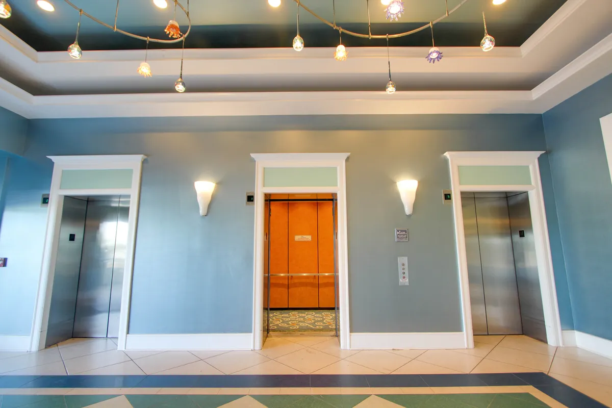Three elevators, one open to reveal a warm wood interior, are set against a decorative ceiling with shell and sea creature ornaments. Soft wall sconces provide ambient lighting, and there is a contrasting tile pattern on the floor.