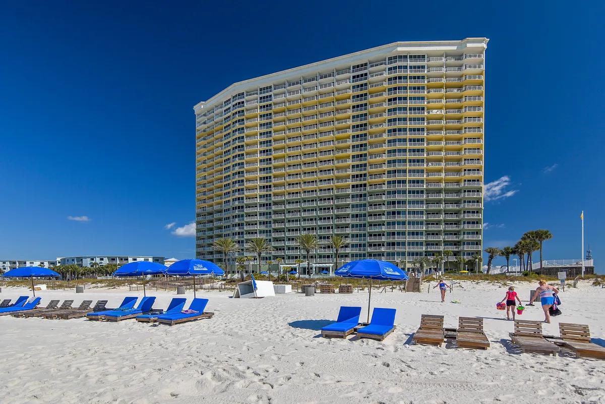 A lively beachfront condominium scene with people enjoying the sandy beach, blue umbrellas, and lounge chairs. The high-rise resort building is visible in the background, with clear blue skies creating an inviting vacation atmosphere.