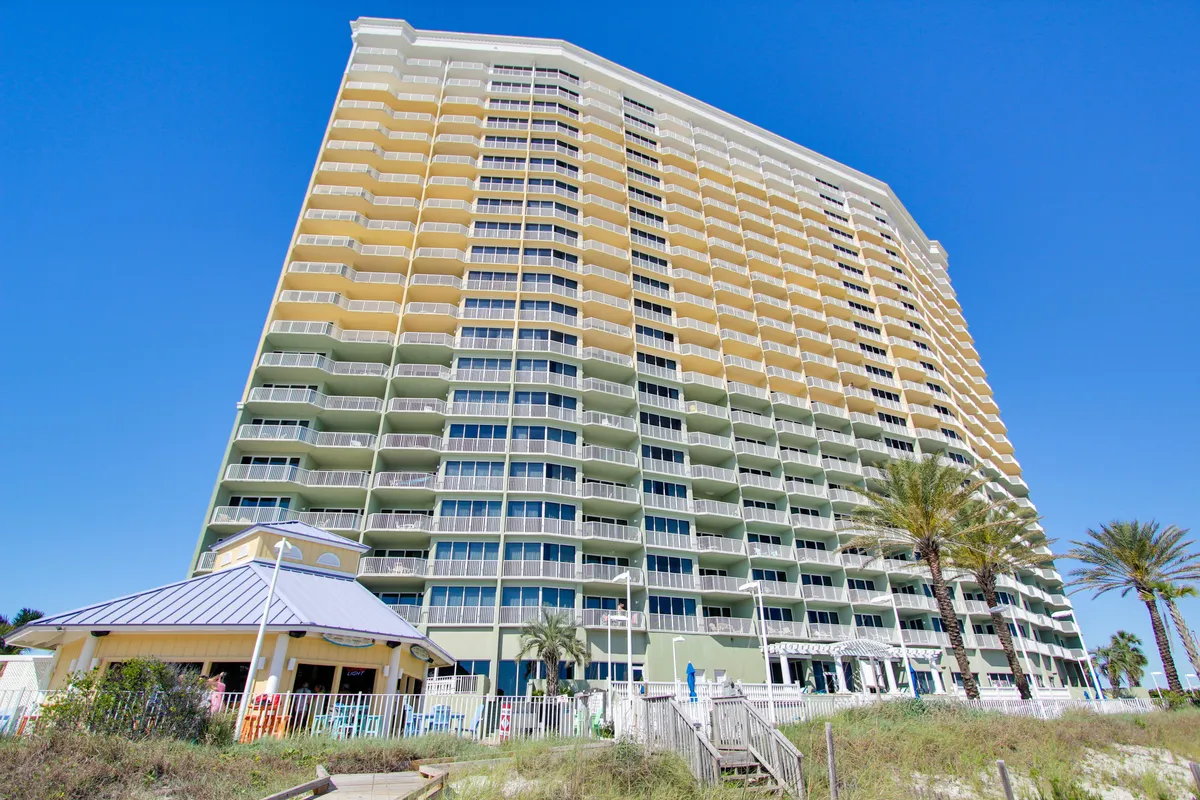 A vibrant beachfront resort scene with a tall condominium building, numerous balconies, a large pool deck with sun loungers, palm trees, and a poolside pavilion or tiki bar. The setting is active yet laid-back, inviting guests to socialize, swim, or relax.