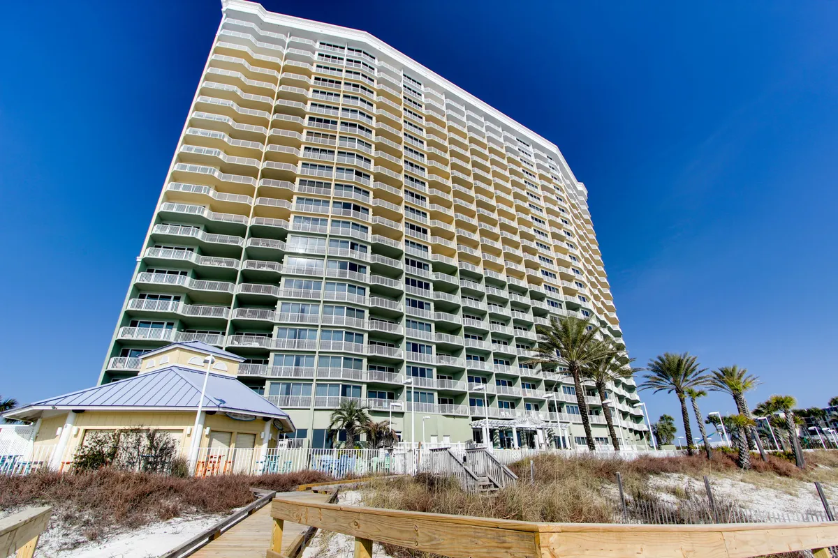 This image features a high-rise condominium building with multiple balconies overlooking a private beach area filled with sand dunes and palm trees. A wooden boardwalk leads directly to the beach, and the clear blue sky enhances the inviting coastal ambiance.