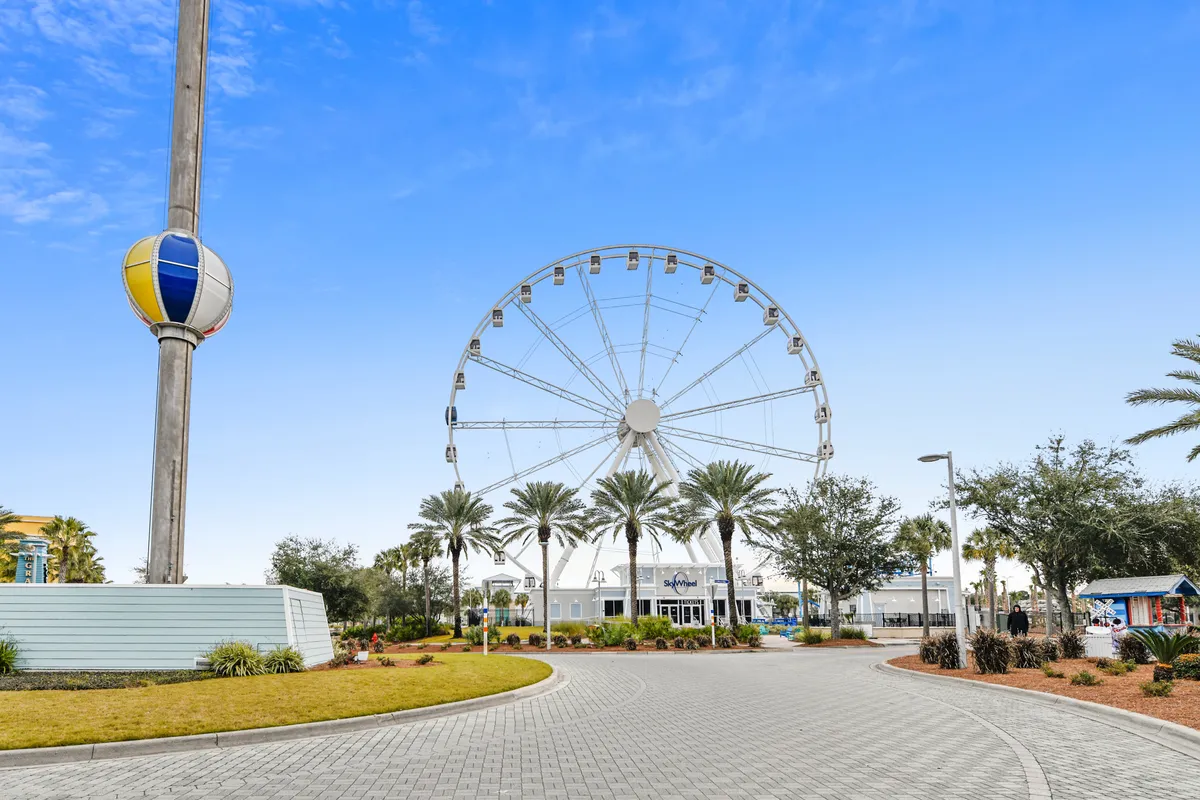 A large Ferris wheel and a colorful balloon-shaped tower are the focal points, complemented by a paved circular walkway and palm trees. The area exudes a lively, cheerful atmosphere, inviting guests to enjoy nearby attractions and seasonal events.