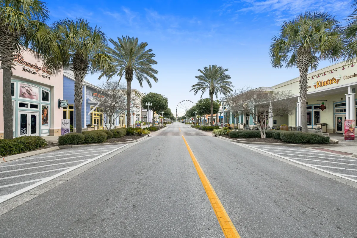 A lively, palm tree-lined street with colorful storefronts and a distant Ferris wheel. The atmosphere is cheerful and welcoming, suggesting a bustling coastal area with shopping, dining, and entertainment options steps away.