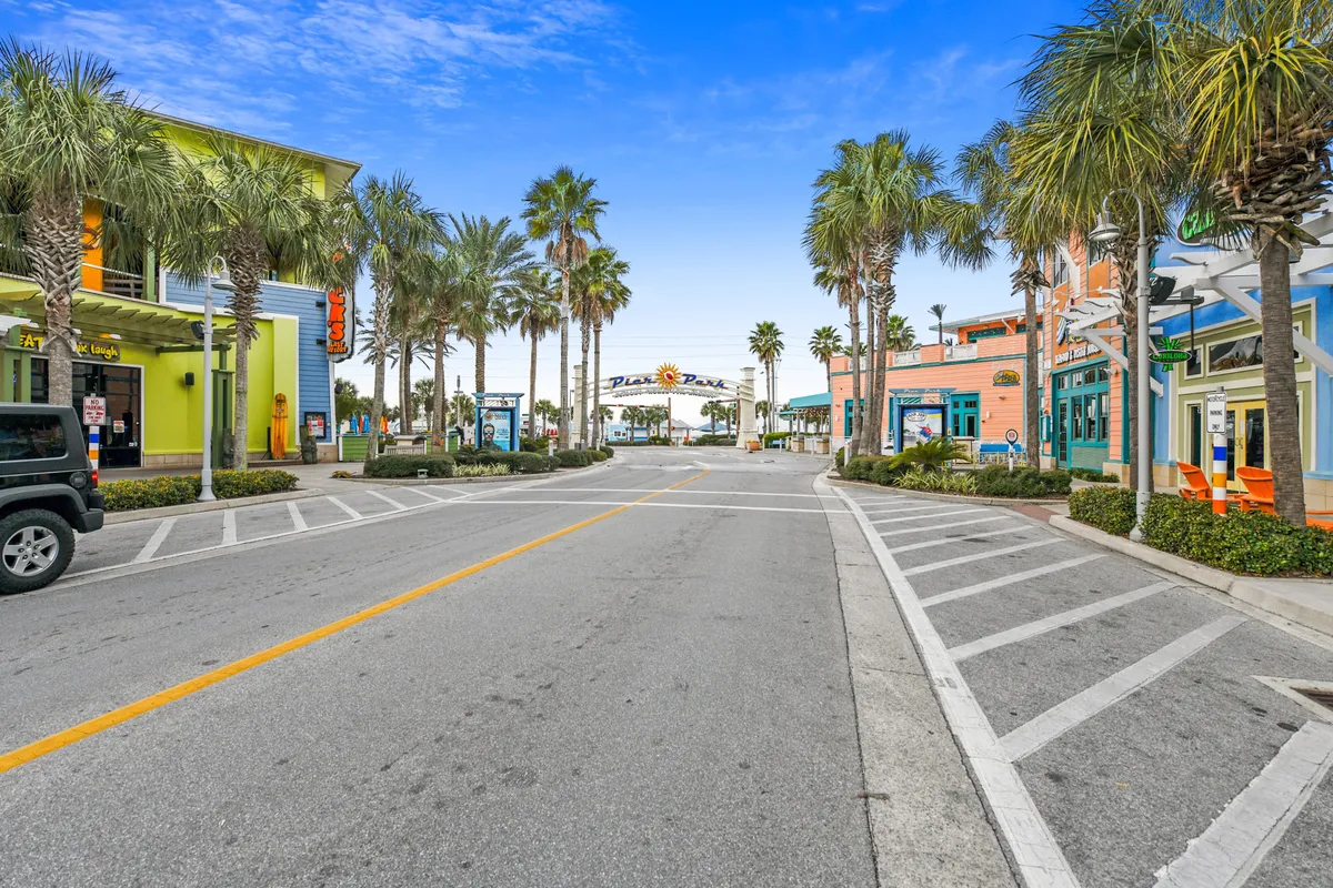 The vibrant entrance to Pier Park is visible in the distance, framed by palm trees, colorful buildings with shops and restaurants, and a clear blue sky. The scene captures the lively, tropical ambiance of the area, inviting exploration and socializing just minutes from the beach.