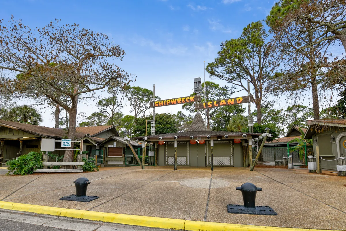 The entrance to Shipwreck Island Water Park is shown with its iconic sign, rustic wooden buildings, and mature trees providing shade. The paved entrance area is empty, setting a scene of anticipation before a day of adventure.